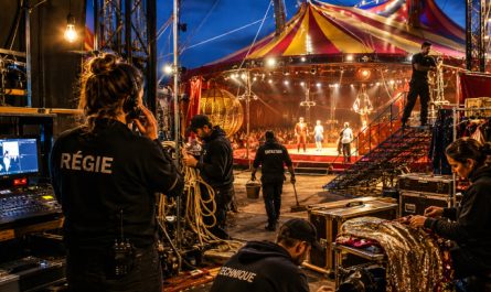 Équipe technique, régie et costumière en coulisses d’un cirque préparant un spectacle sous un grand chapiteau illuminé